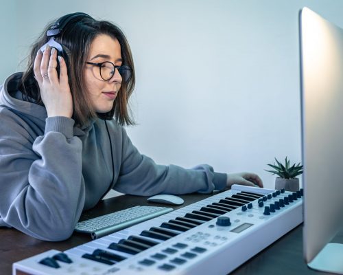 A young woman creates music using a musical keyboard and a computer. A young woman musician at workplace, creates music using musical keyboard and computer, freelance, musical creativity.